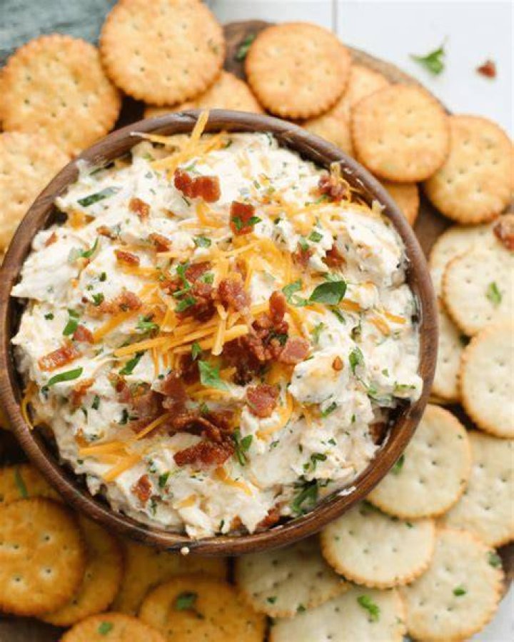 Bowl of dip topped with artichokes and sun dried tomatoes served with vegetables and pretzels.